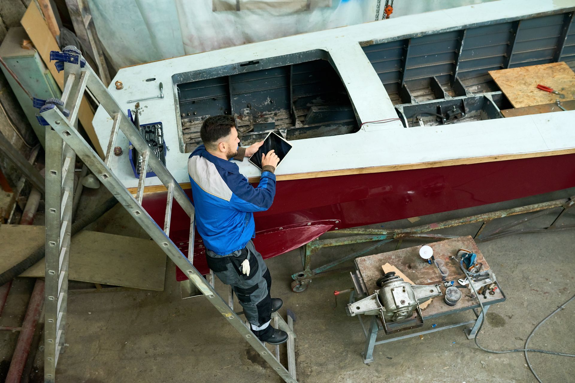 Man on ladder inspects boat hull with tablet, tools on nearby table.