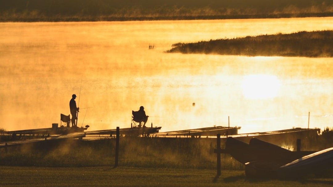 Two people are fishing on a dock at sunset.