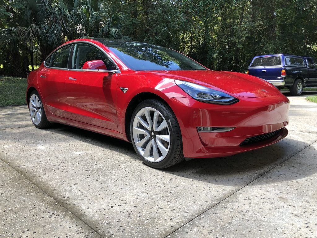 A red tesla model 3 is parked in a driveway.