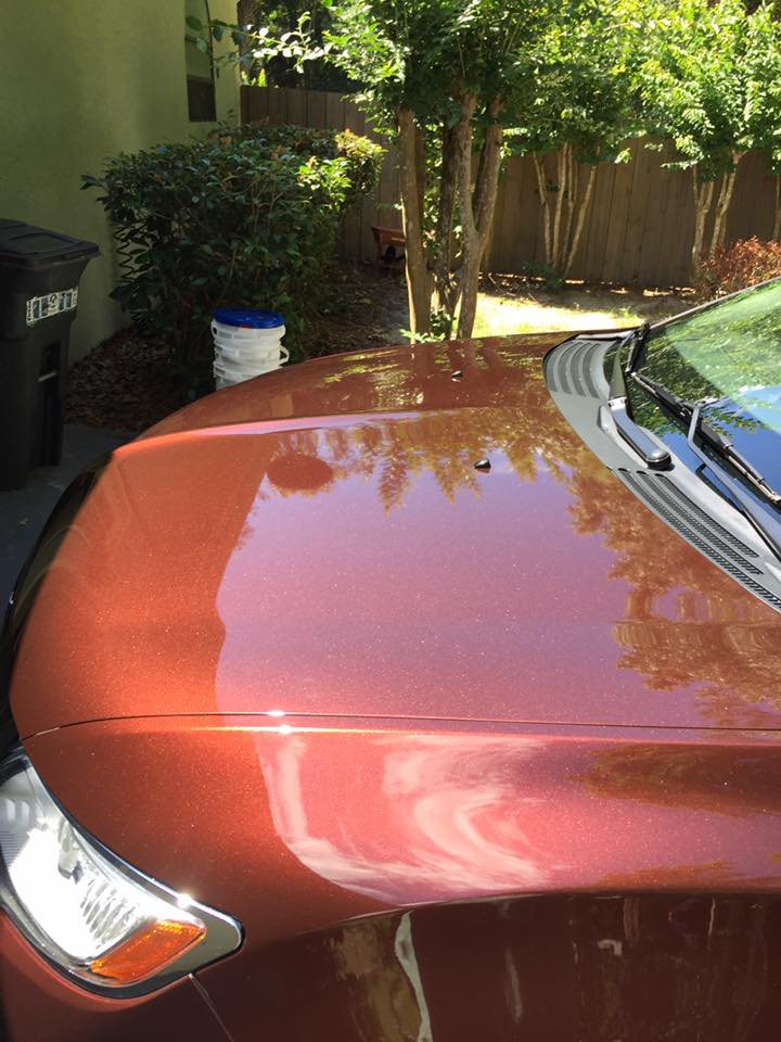 A red car is parked in a driveway next to a trash can.