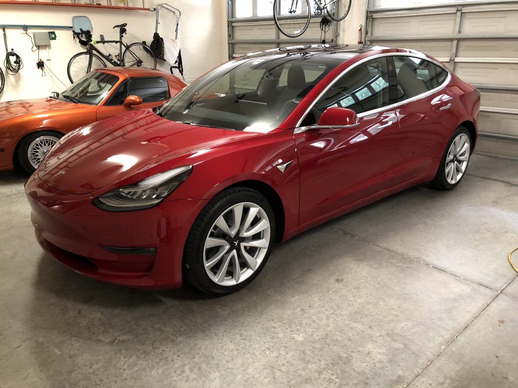 A red tesla model 3 is parked in a garage next to a corvette.