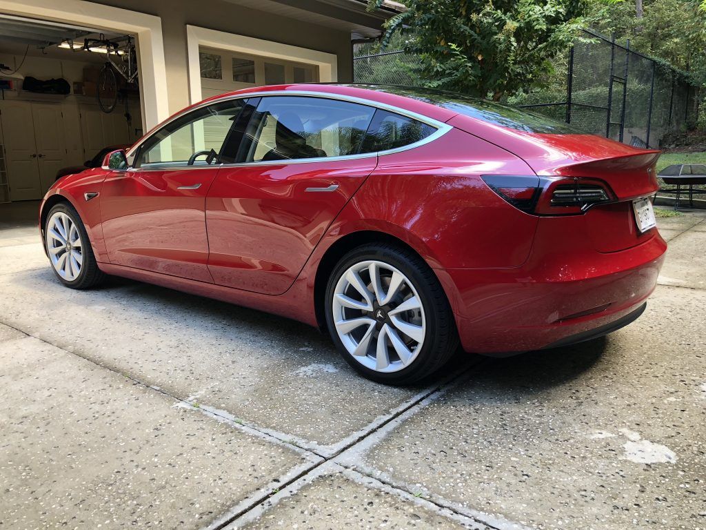 A red tesla model 3 is parked in a driveway in front of a garage.