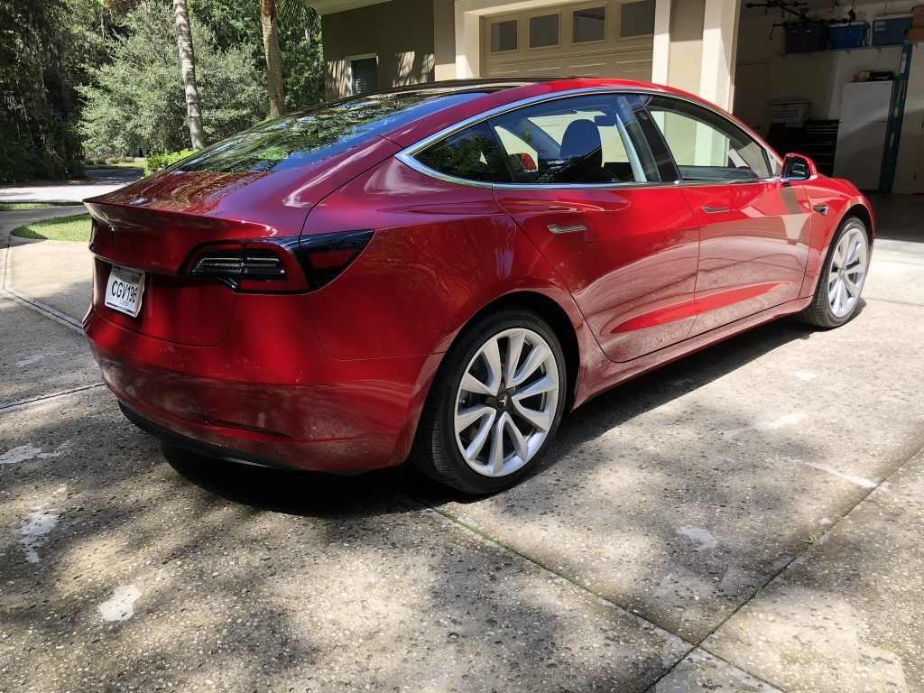 A red tesla model 3 is parked in front of a garage.