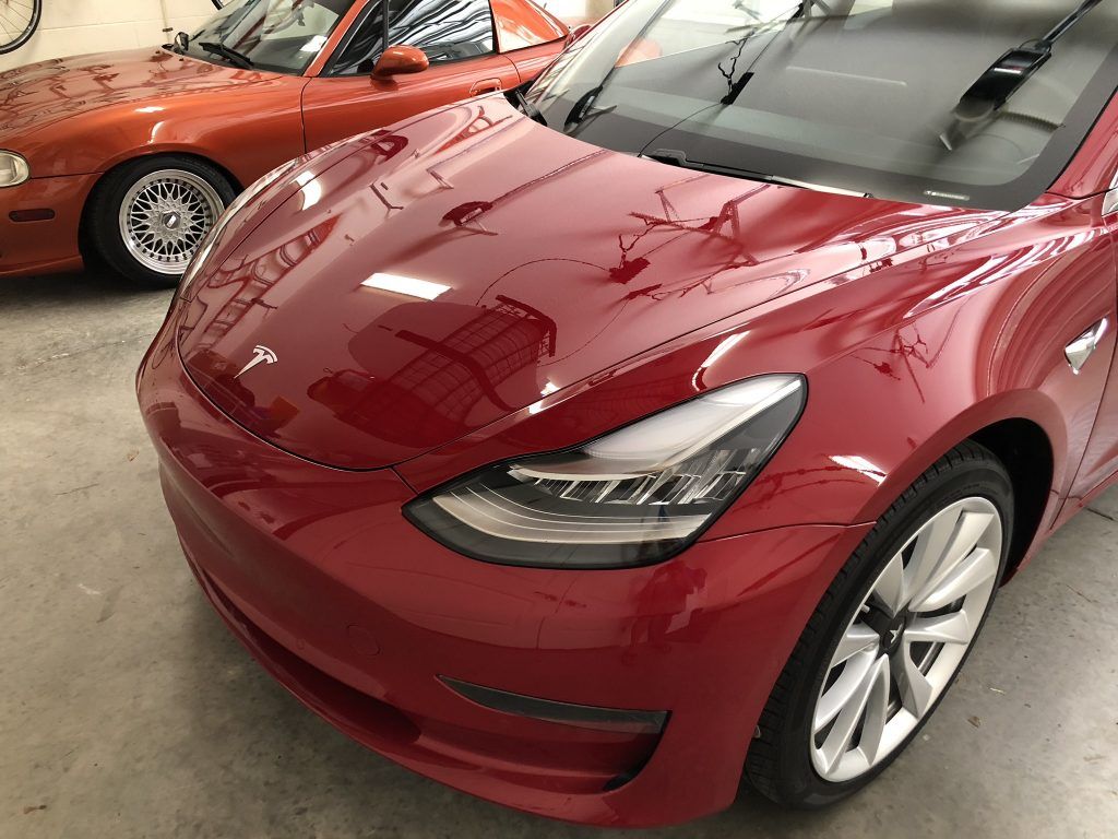 A red tesla model 3 is parked next to another red car in a garage.