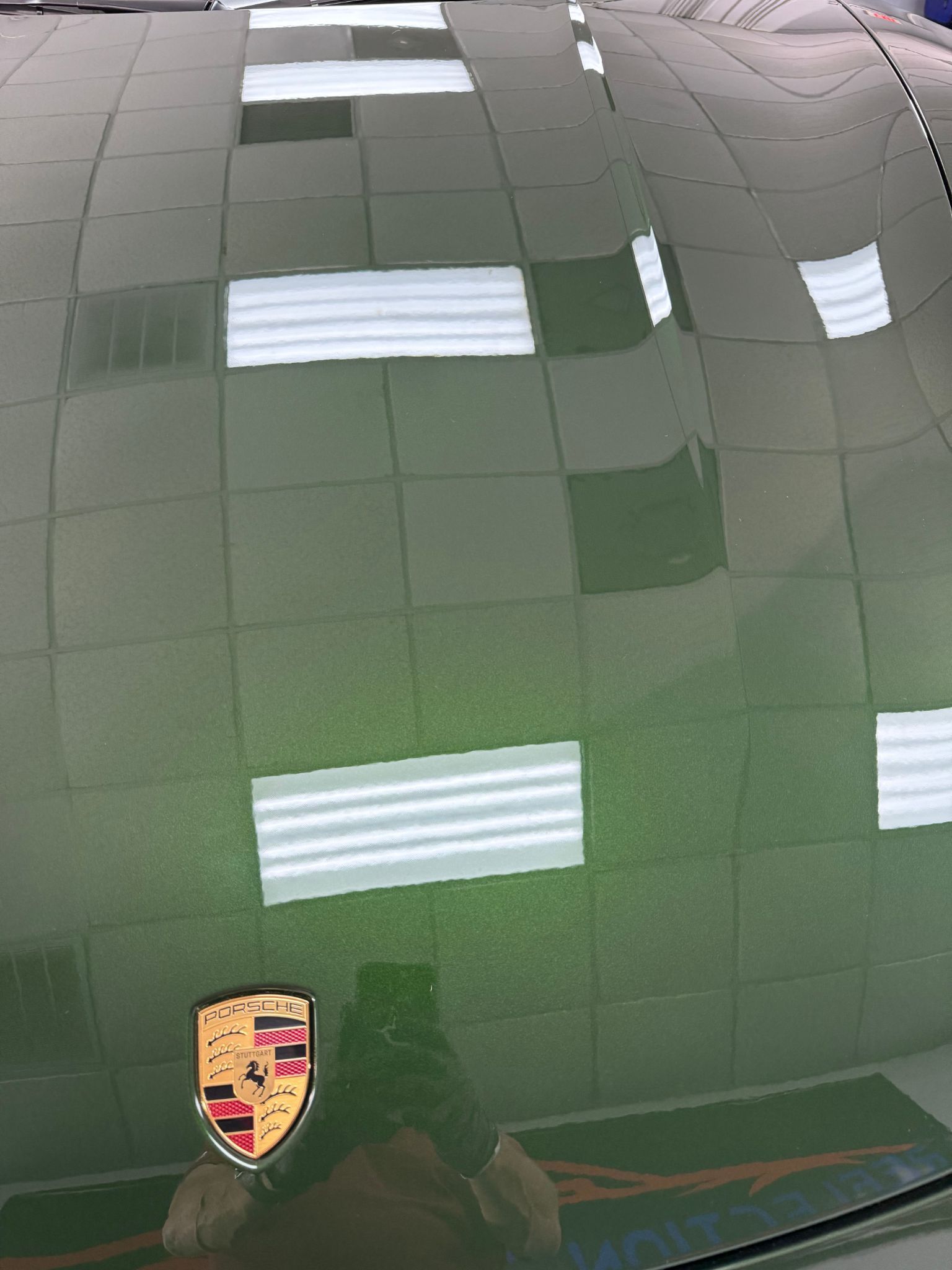 Green Porsche hood reflecting overhead lights; car emblem visible.