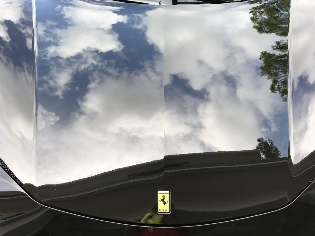 The hood of a black ferrari is reflecting the sky and clouds.