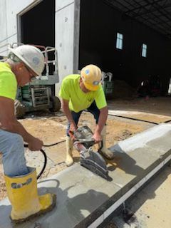 Two construction workers cutting concrete; one operates saw, other holds hose. Both wear safety gear outside a building.