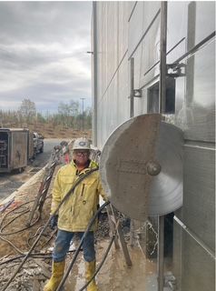 Construction worker in yellow jacket operating wall saw, cutting into building exterior.