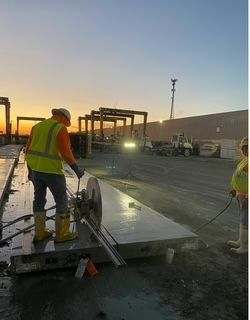 Construction worker cutting concrete slab with a circular saw outdoors, sunset in the background.