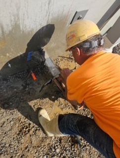 Construction worker in orange shirt and hard hat uses a tool near a concrete surface outdoors.