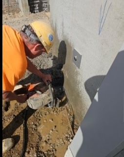 Construction worker using a jackhammer on concrete near a wall, wearing a hard hat and orange shirt.