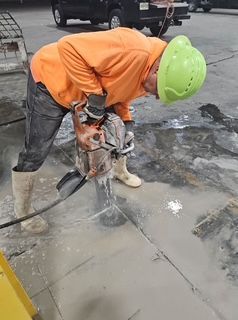 Worker in orange shirt and green helmet, using a jackhammer to break up concrete floor.
