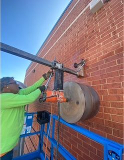 Worker drilling a large hole in a brick wall from a lift, wearing a green shirt, using an orange drill.