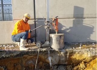 Construction worker using a coring drill on a concrete wall. Outdoor setting; worker wearing safety gear.
