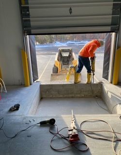 Man in orange jacket uses jackhammer, digging inside a warehouse loading dock, skid steer in background.