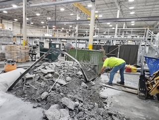 Man in neon shirt breaking concrete with a tool in an industrial warehouse. Debris pile, machinery.