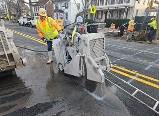 Construction worker operating a road saw on asphalt, another worker nearby. Yellow safety vests, street setting.