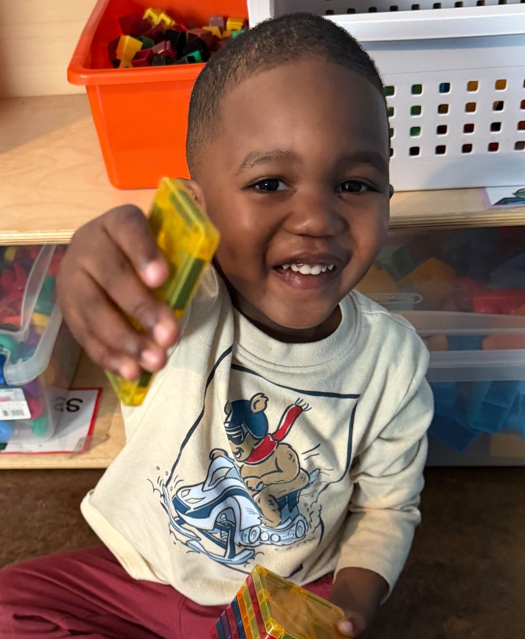 Young child smiling, holding yellow magnetic tiles; indoors near toy storage.
