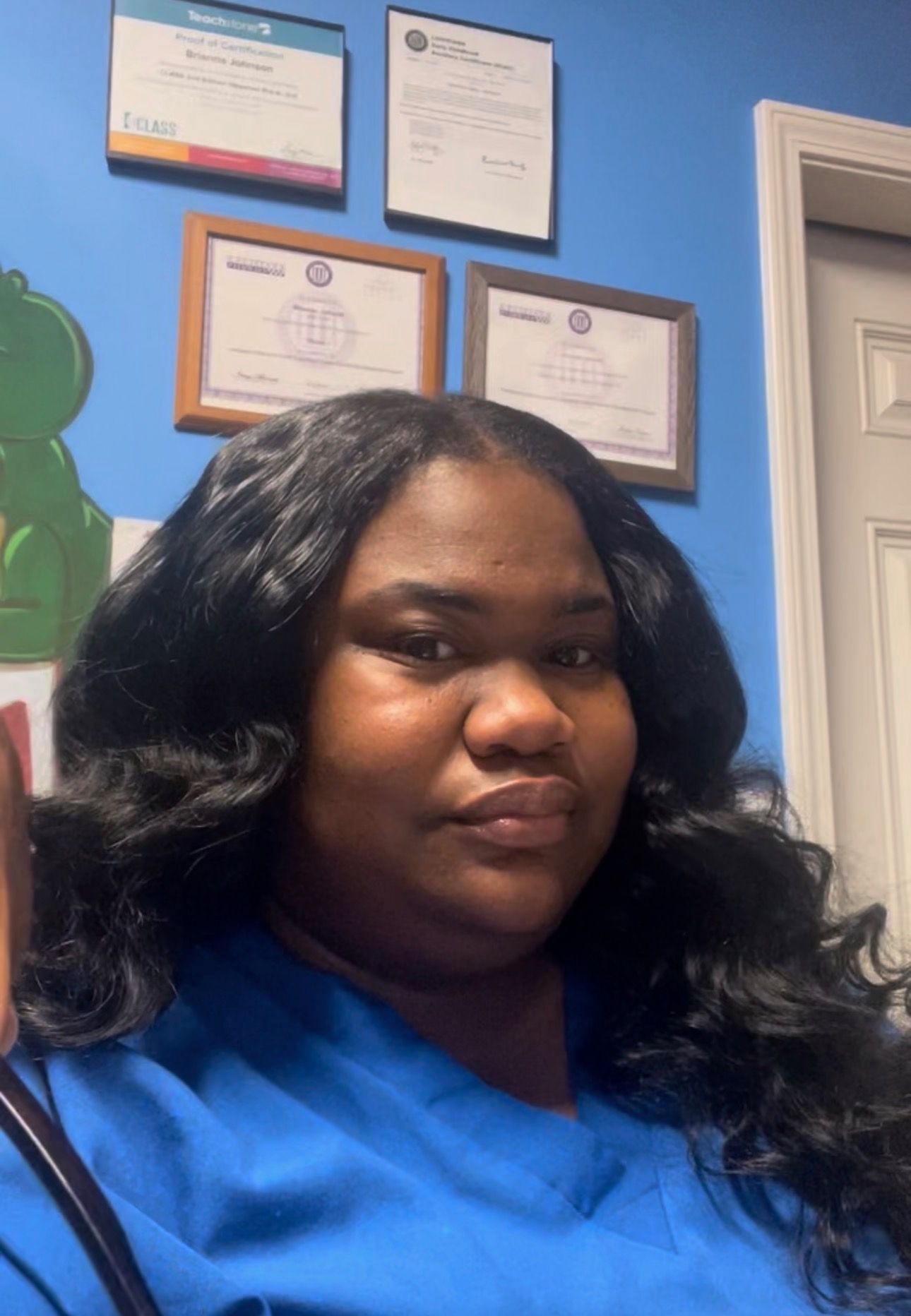 Woman with dark hair in a blue shirt, standing in front of a blue wall with framed certificates.