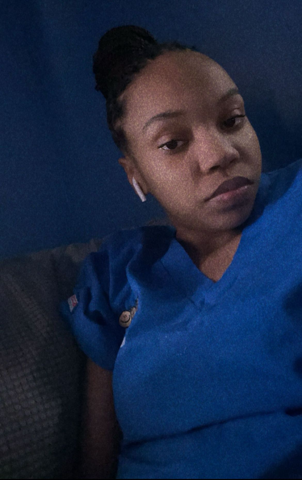 Person in blue scrubs with hair in a bun wearing earbuds, looking toward the camera.