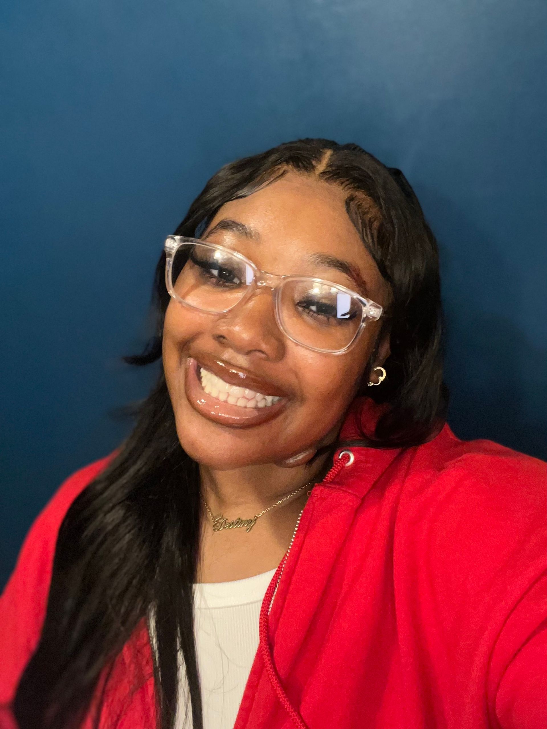 Woman smiling, wearing clear glasses and red jacket, against a blue background.