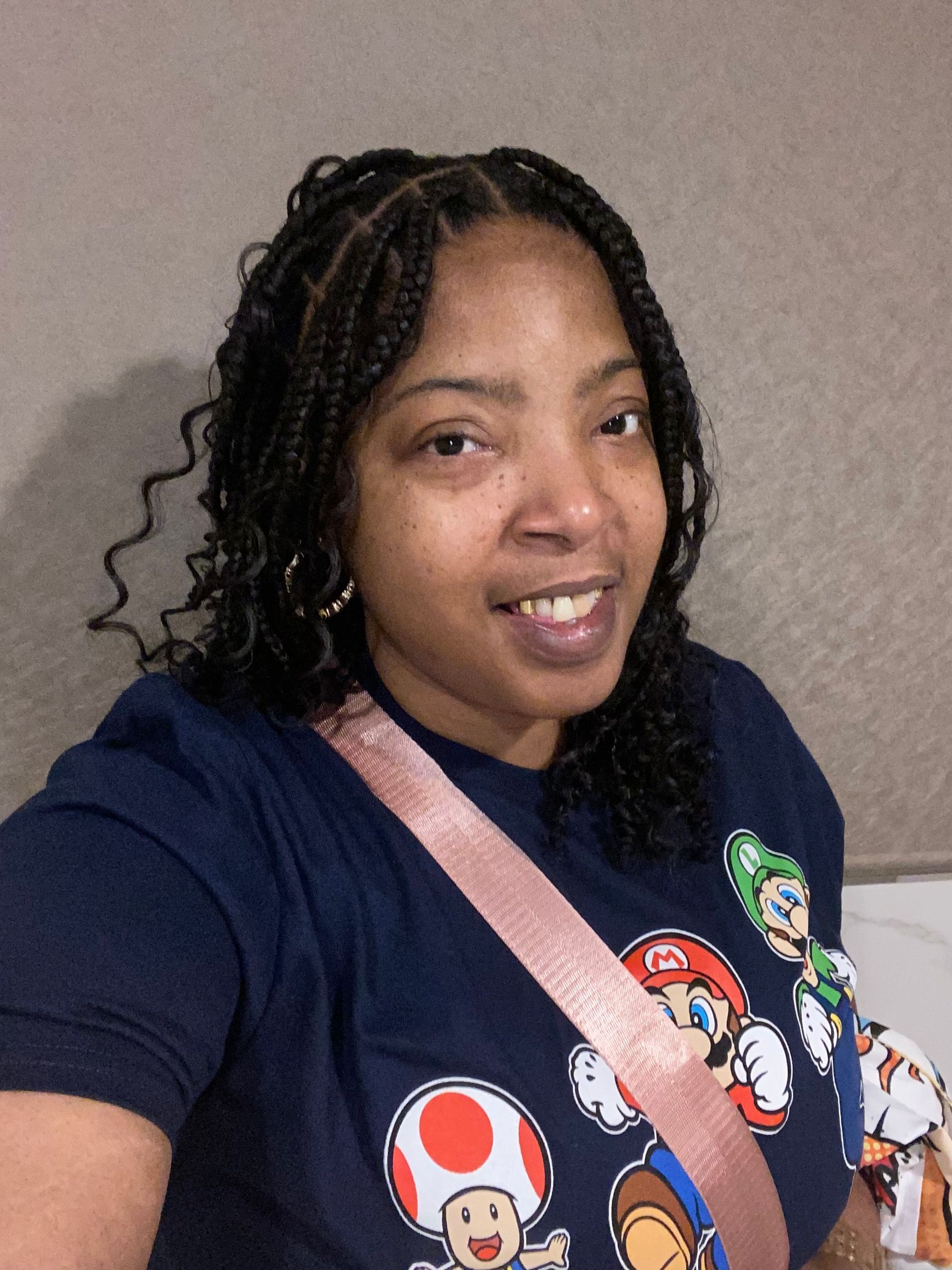 Woman with braided hair, wearing a navy Mario t-shirt, smiles. Pink purse strap across the chest.