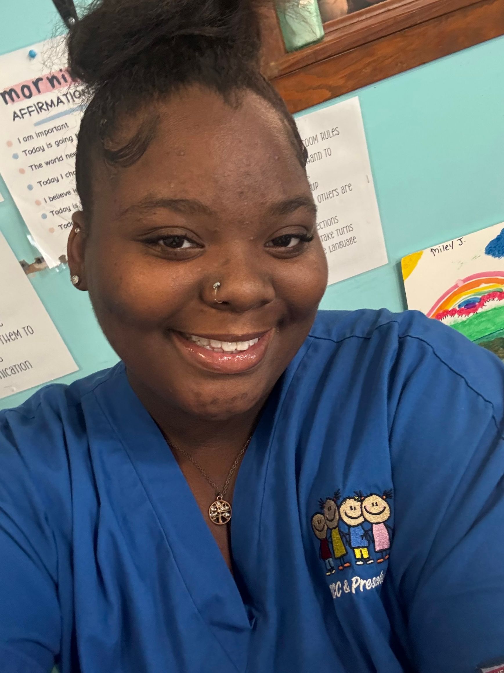 Woman in blue scrubs smiles, with hair up. Rainbow drawing and a list are in the background.