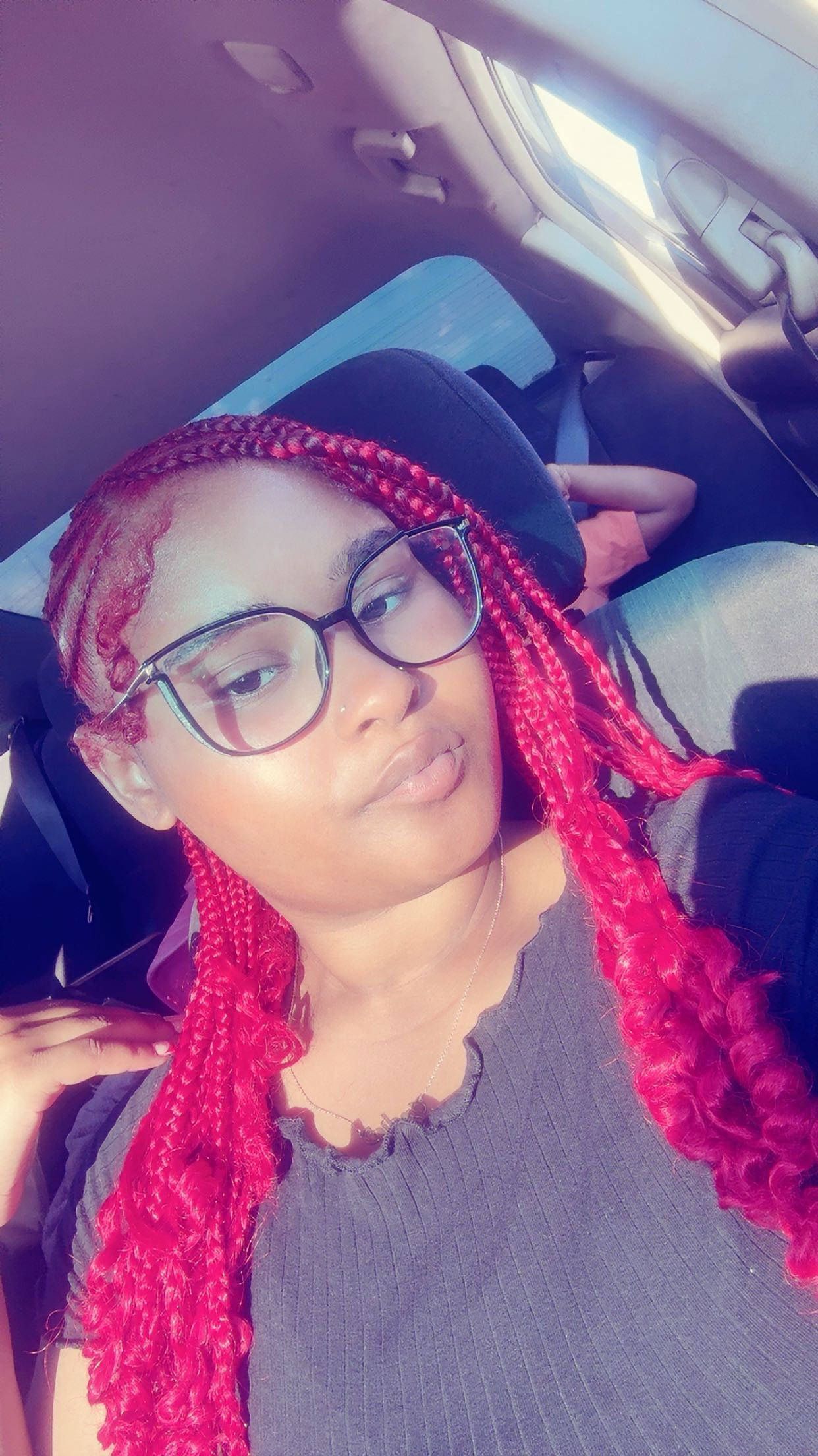 Person with red braided hair and glasses, sitting in a car, looking at the camera.