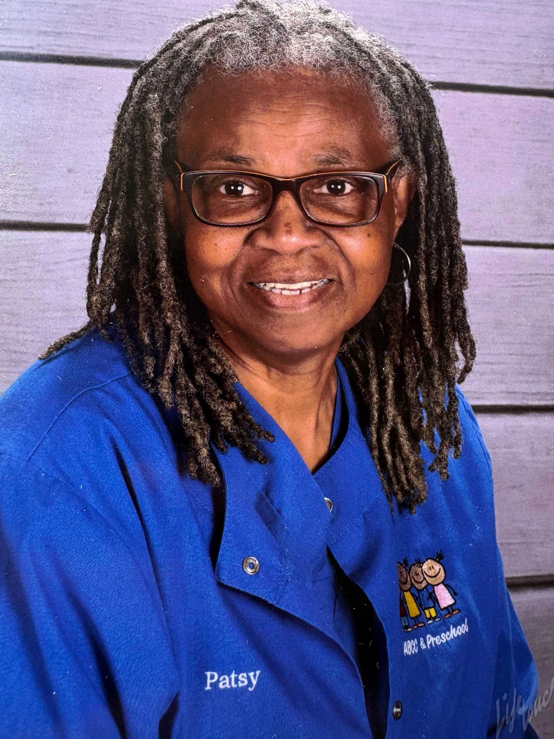 Woman with glasses and dreadlocks wearing a blue shirt with a smiling expression.