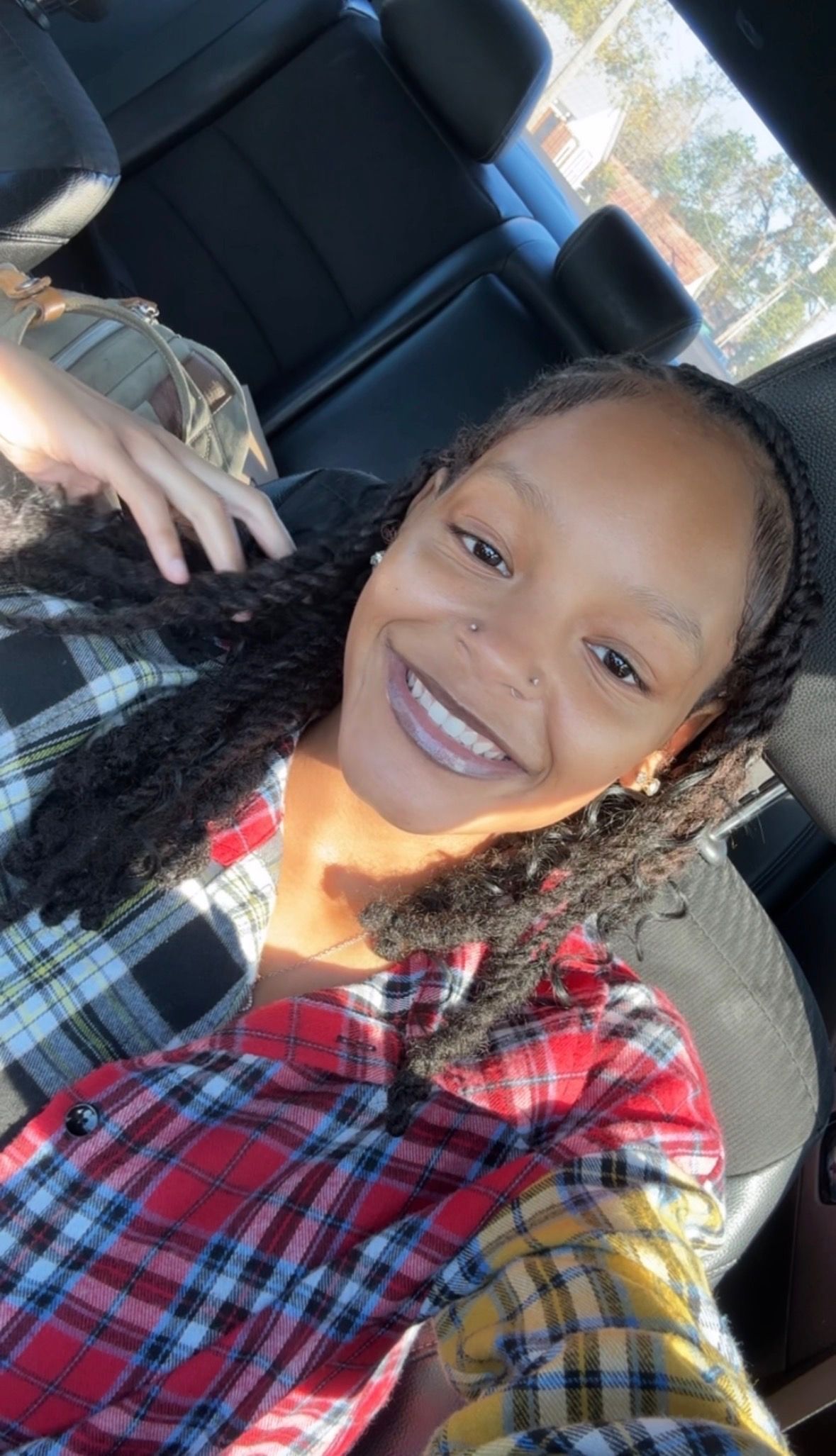 Person with dark braids smiles in a car, wearing a plaid shirt. Sunlight illuminates the face.