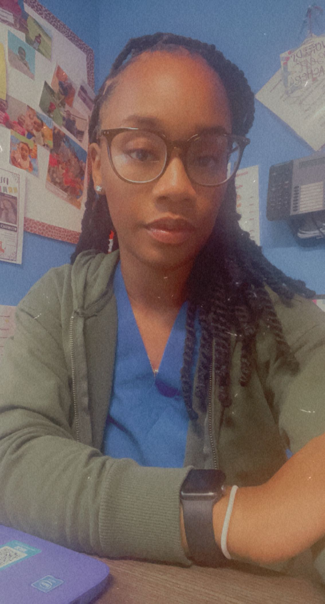 Woman with glasses, dark hair, wearing green jacket and blue top, sitting at a desk with an Apple watch on.