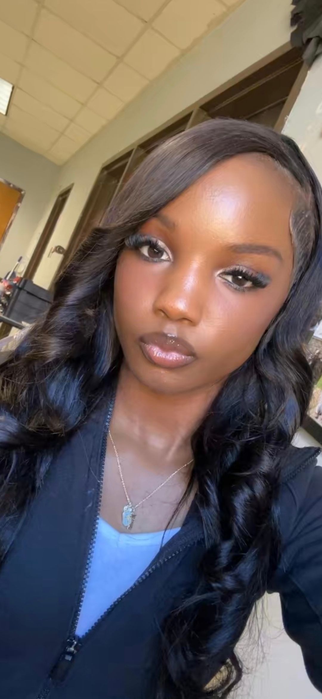 A person with long, wavy black hair in a black zipped jacket takes a selfie. She is in a room.