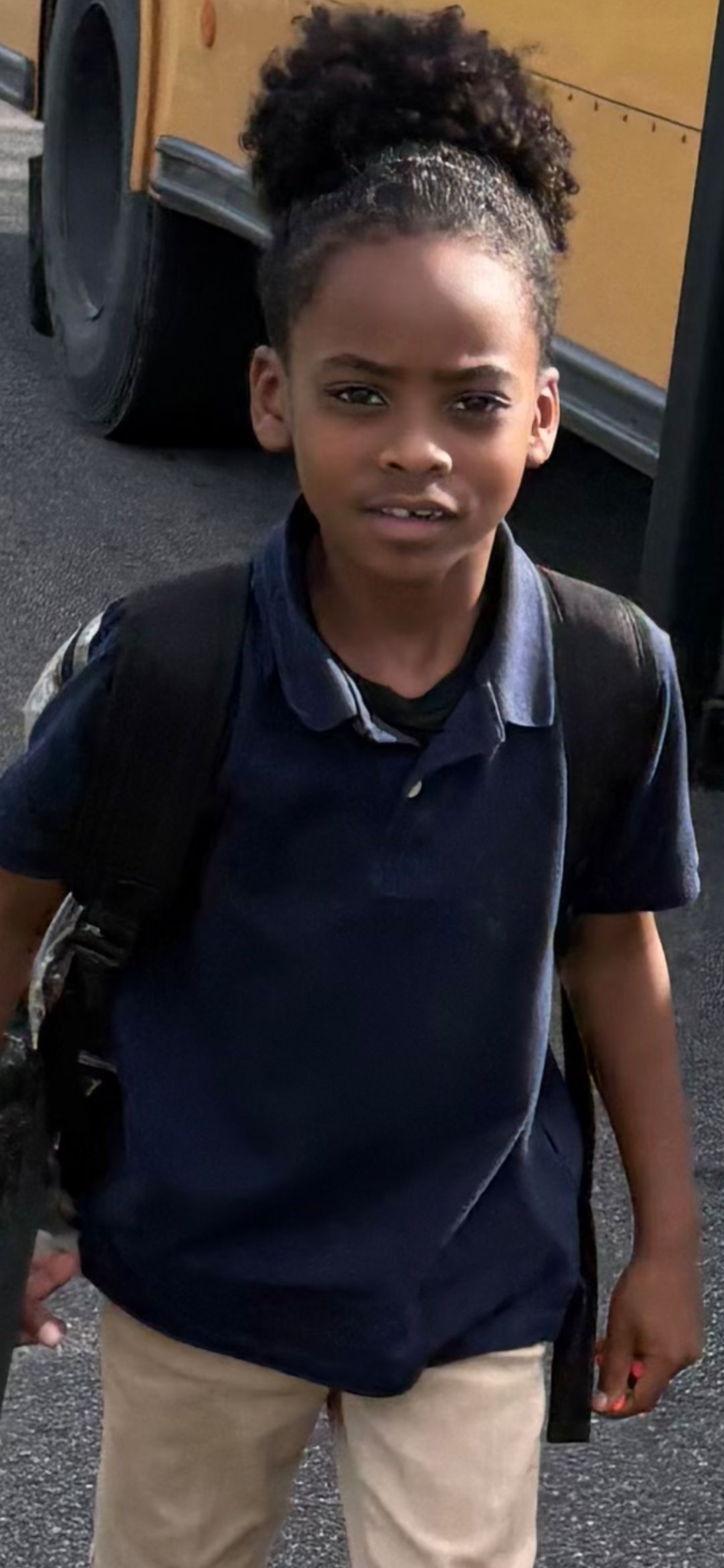 A person with hair pulled up wearing a navy polo shirt and tan pants, with a backpack.