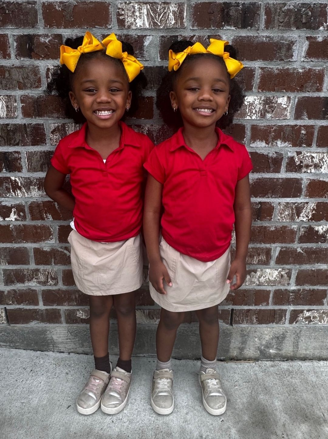 Two girls in school uniforms smile in front of a brick wall. Each wears a red shirt, khaki skirt, and yellow bow. Pre K