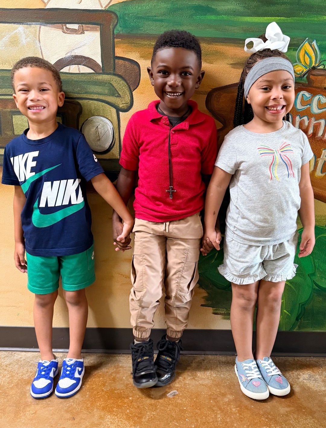 Three children holding hands smiling, standing against a painted wall with a truck, wearing casual clothes.