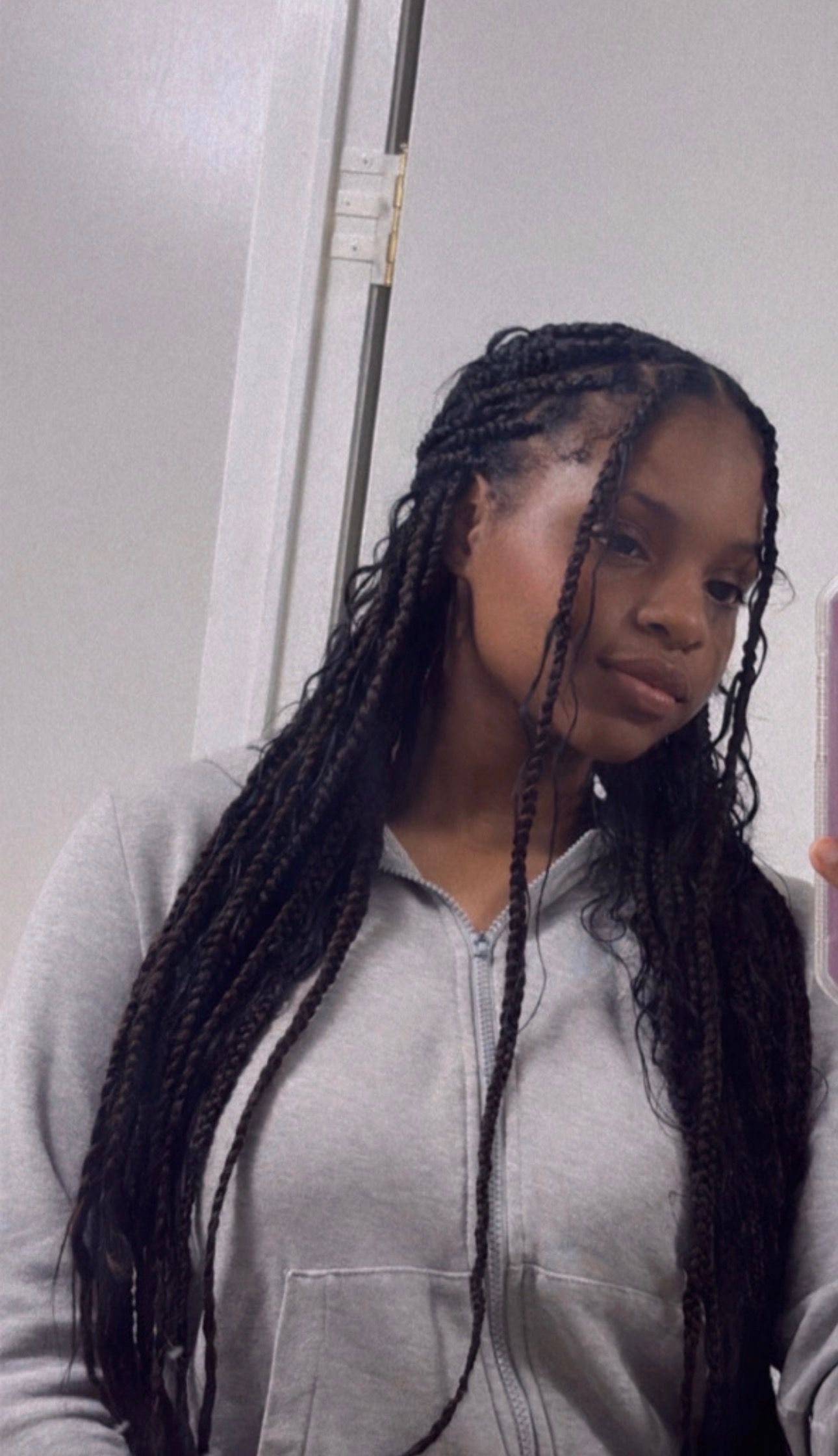 Woman with long braids holding phone, looking at the camera in a gray hoodie indoors.