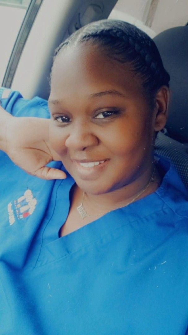 Woman in blue scrubs smiles, head resting on hand, in car.