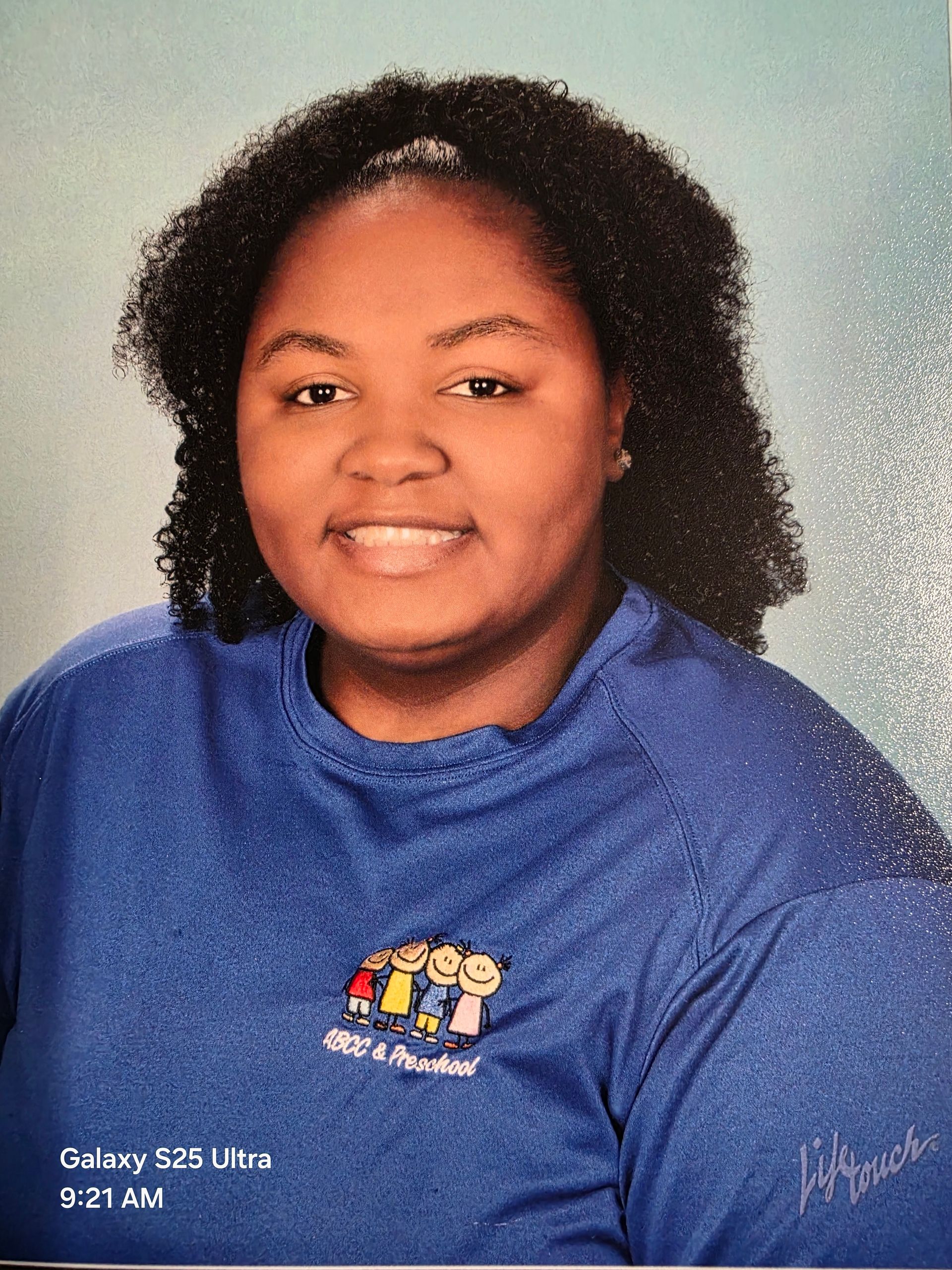 Smiling person with curly dark hair wearing a blue shirt with a childcare logo.