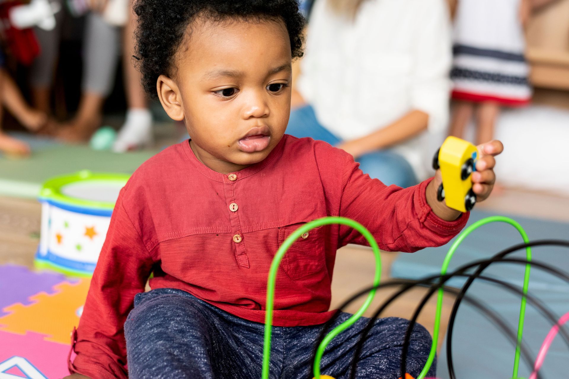 Boy playing with a bead maze toy in a playroom, red shirt, other children in background.