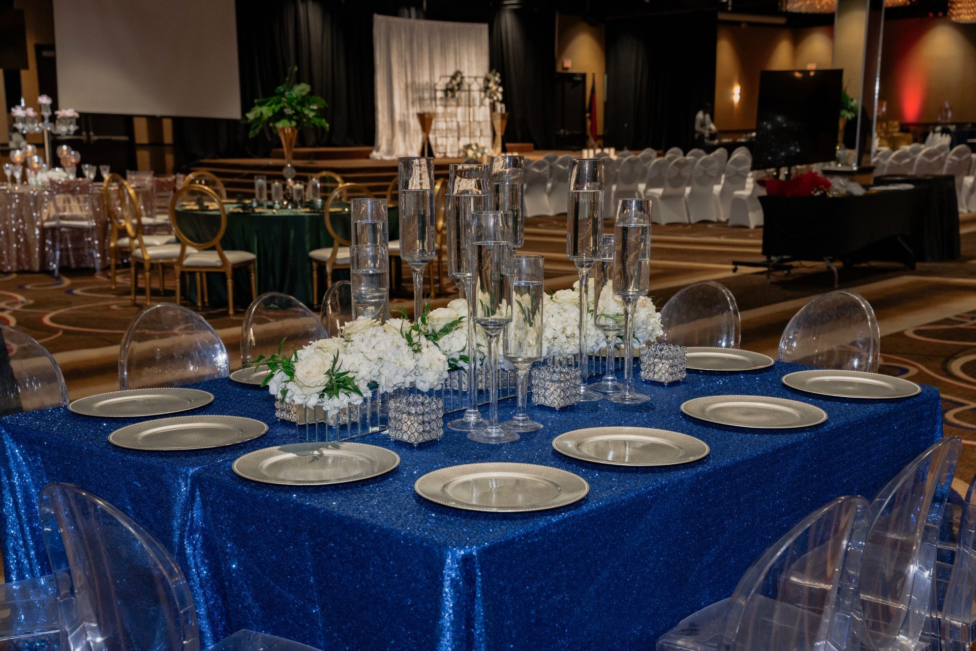 A table set for a wedding reception with a blue tablecloth and clear chairs.