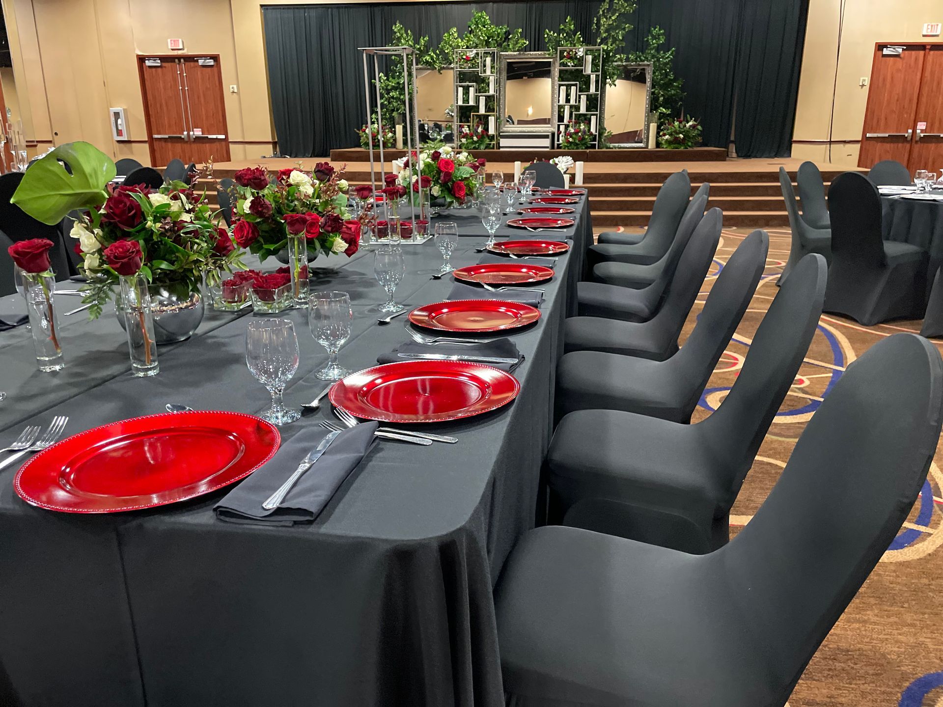 A long table with red plates , glasses , and vases of flowers.