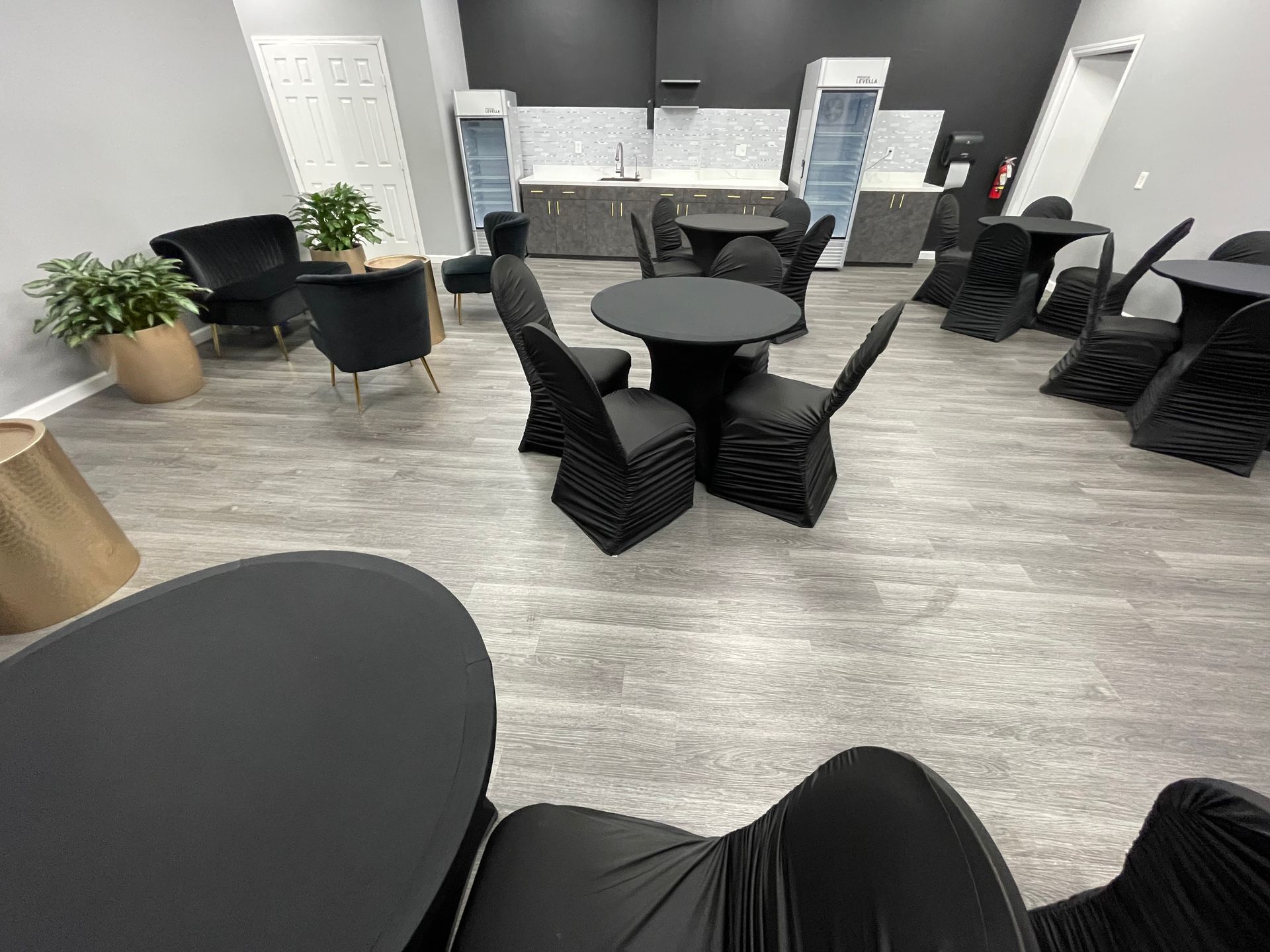 The Collective Lounge area with black tables, chairs, and cabinets. Plants, light gray flooring, and a white backsplash are visible.