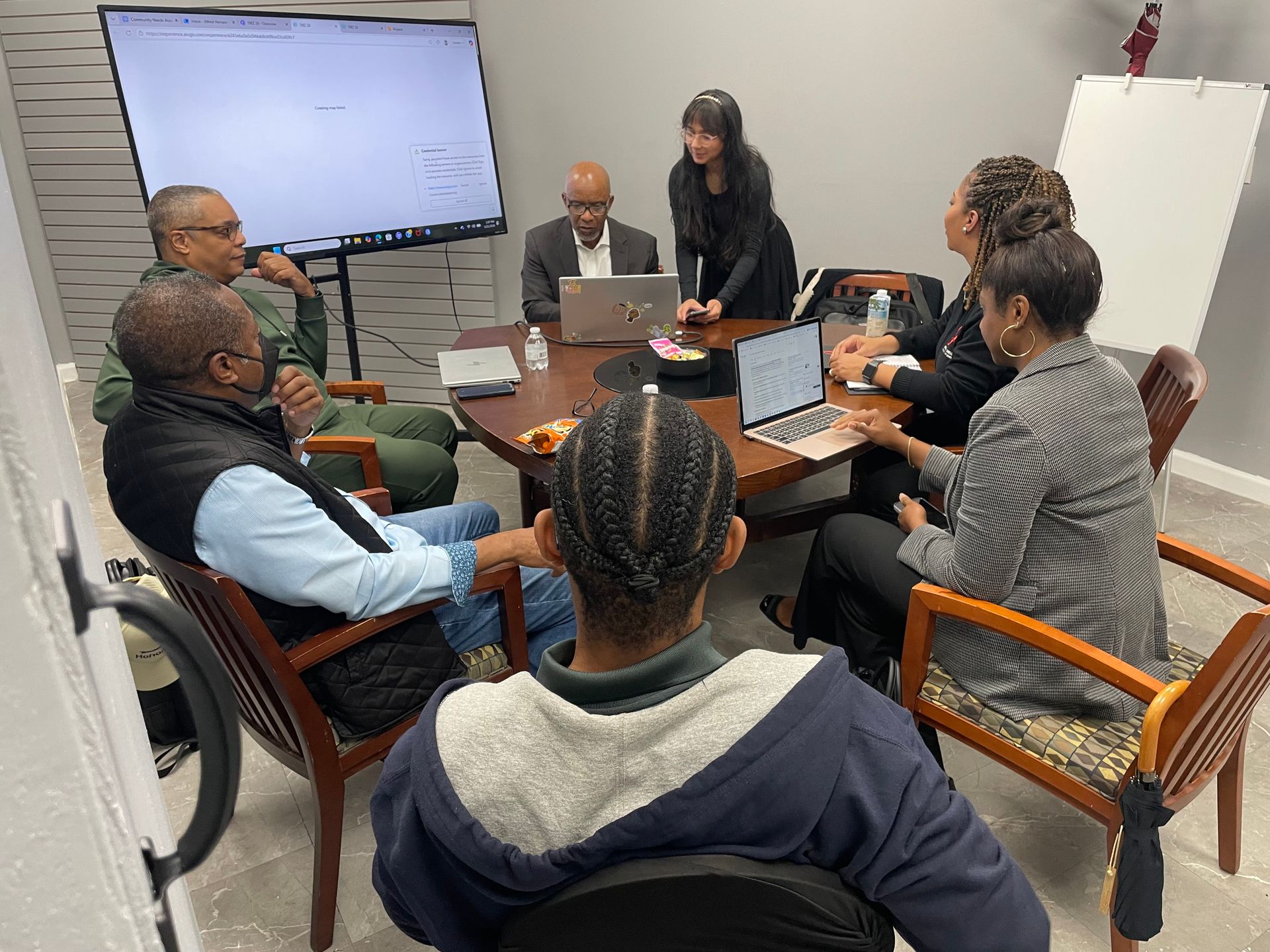 People in a meeting around a table, some using laptops. A large screen and whiteboard are visible in the room.