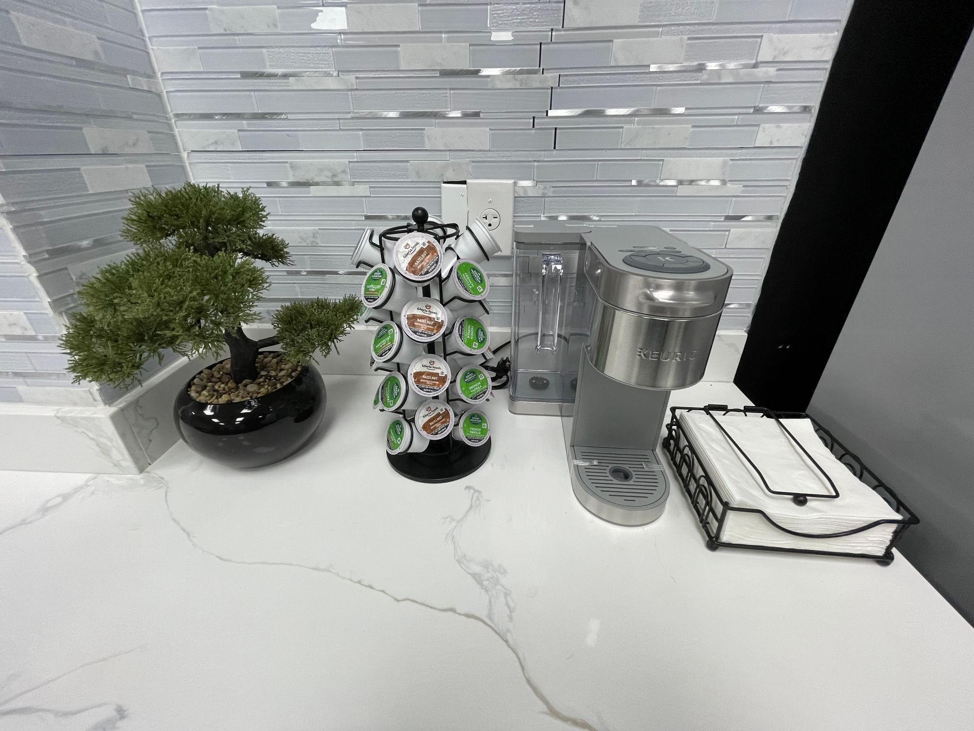 Coffee station with a Keurig, K-cup carousel, bonsai tree, and napkin holder on a white countertop with a tiled backsplash.