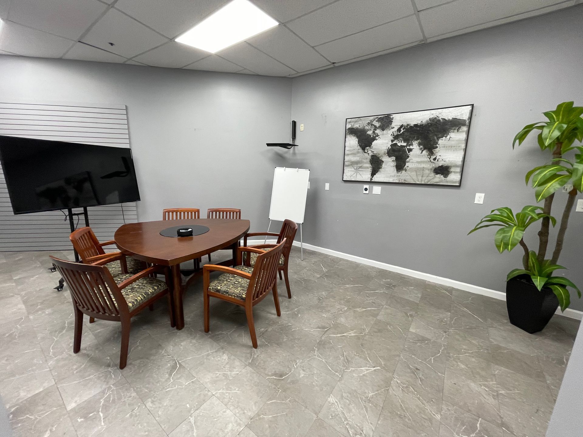 Meeting room with a wooden table, chairs, large screen, whiteboard, world map art, and a potted plant.