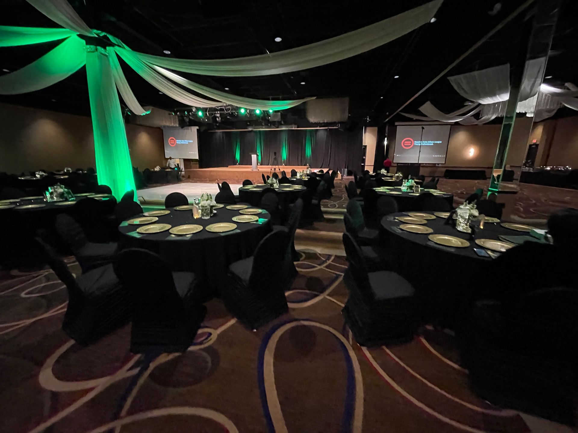 Ballroom set for an event with round tables, black linens, and a stage with green lighting and decor.