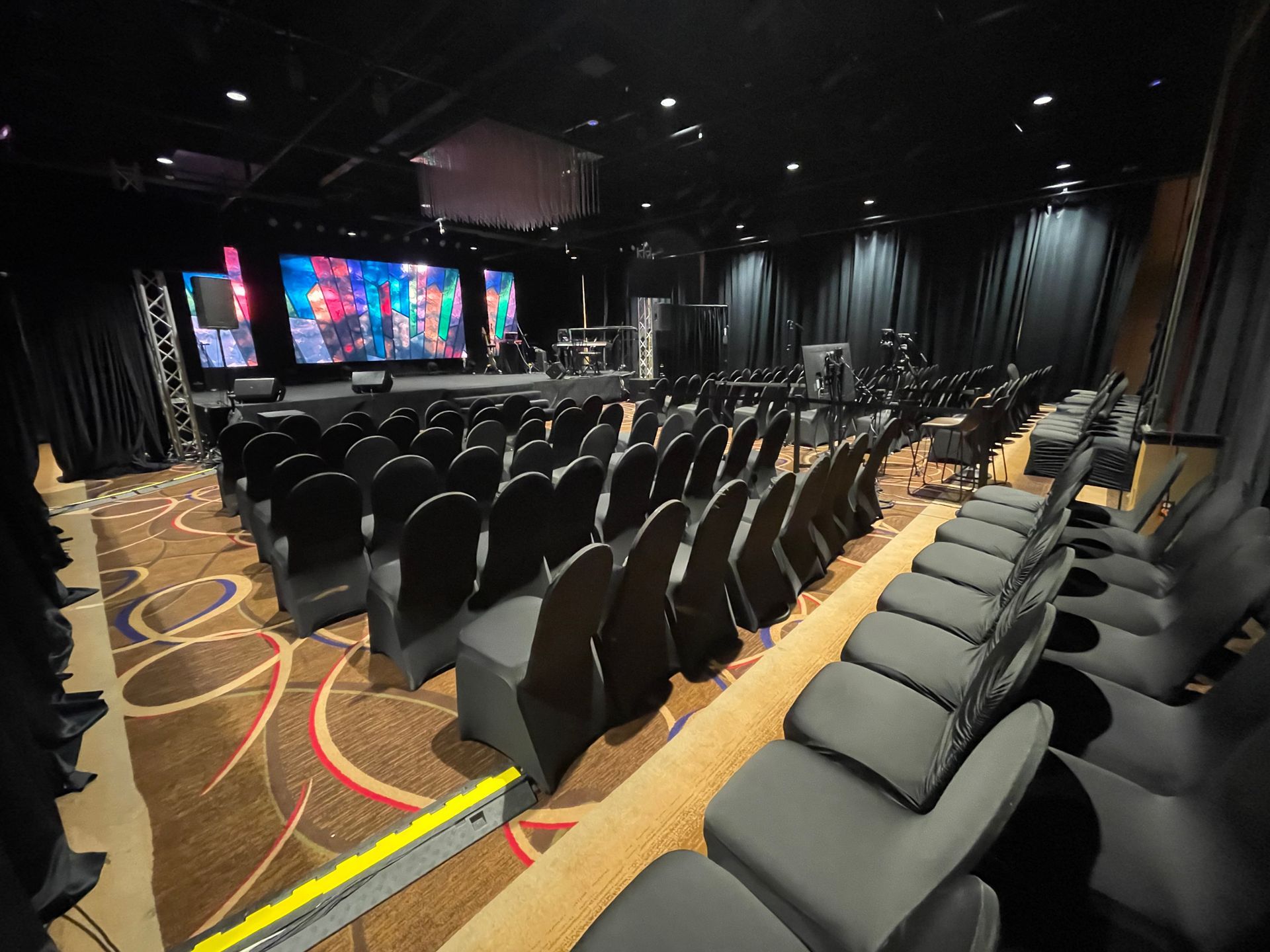 Auditorium with rows of black chairs facing a stage with screens and instruments. Dark setting.