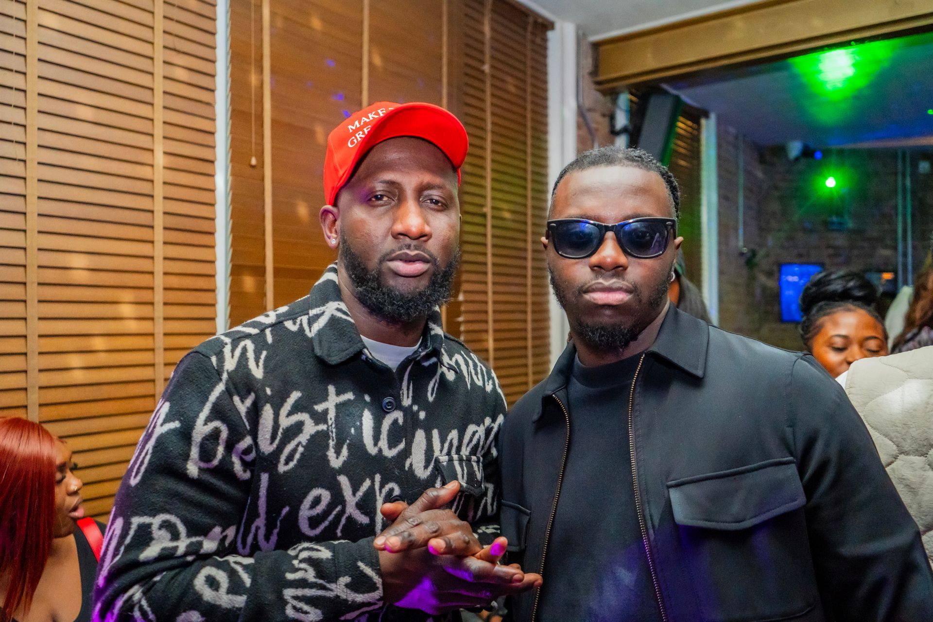 Two men are standing next to each other in a room . one of the men is wearing sunglasses and a red hat.