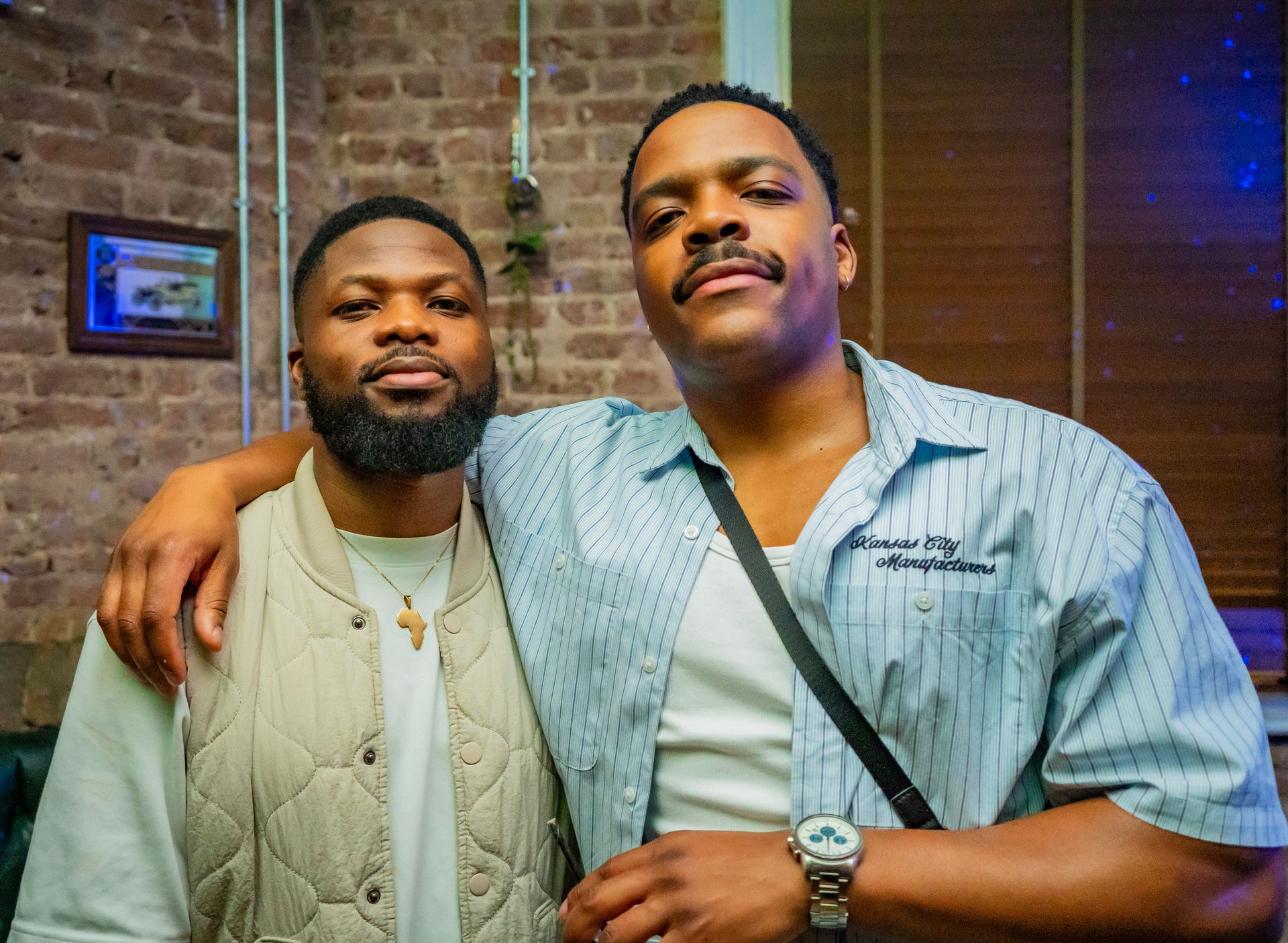 Two men are posing for a picture together in front of a brick wall.