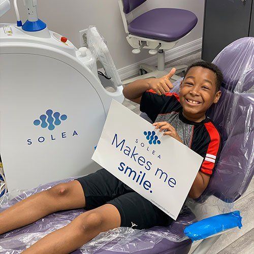A young boy is sitting in a dental chair holding a sign that says `` makes me smile ''.